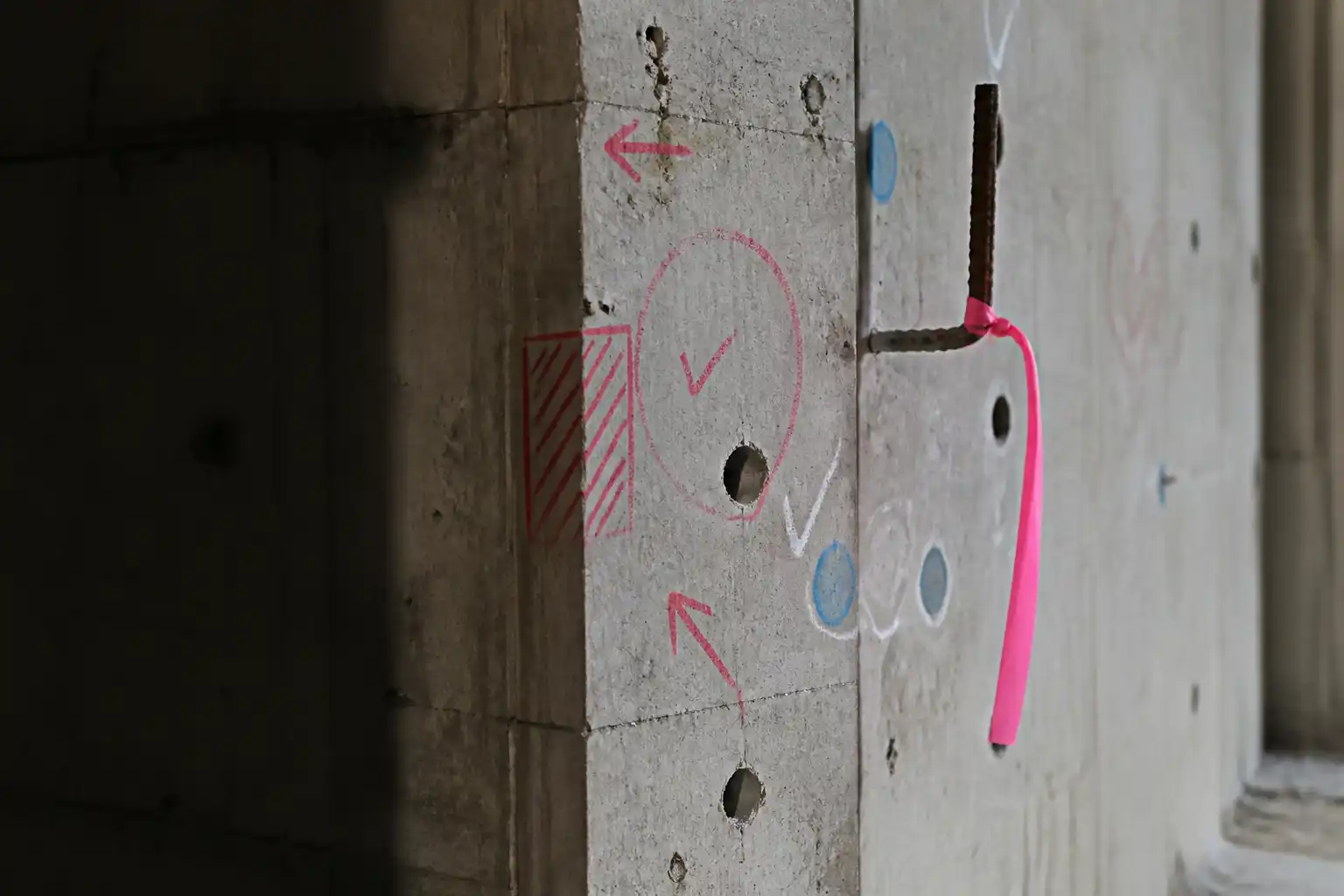 A concrete column marked up by hand in crayon and chalk, with flagging tape tied to protruding rebar